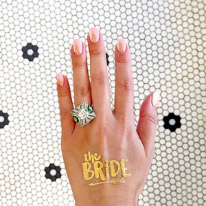 May include: A hand with gold glitter nail polish and a large diamond ring with green stones. The hand has a gold temporary tattoo that says "the bride" with an arrow.