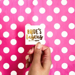 May include: White square temporary tattoo with gold lettering that reads "Bride's besties" with a small gold arrow underneath.