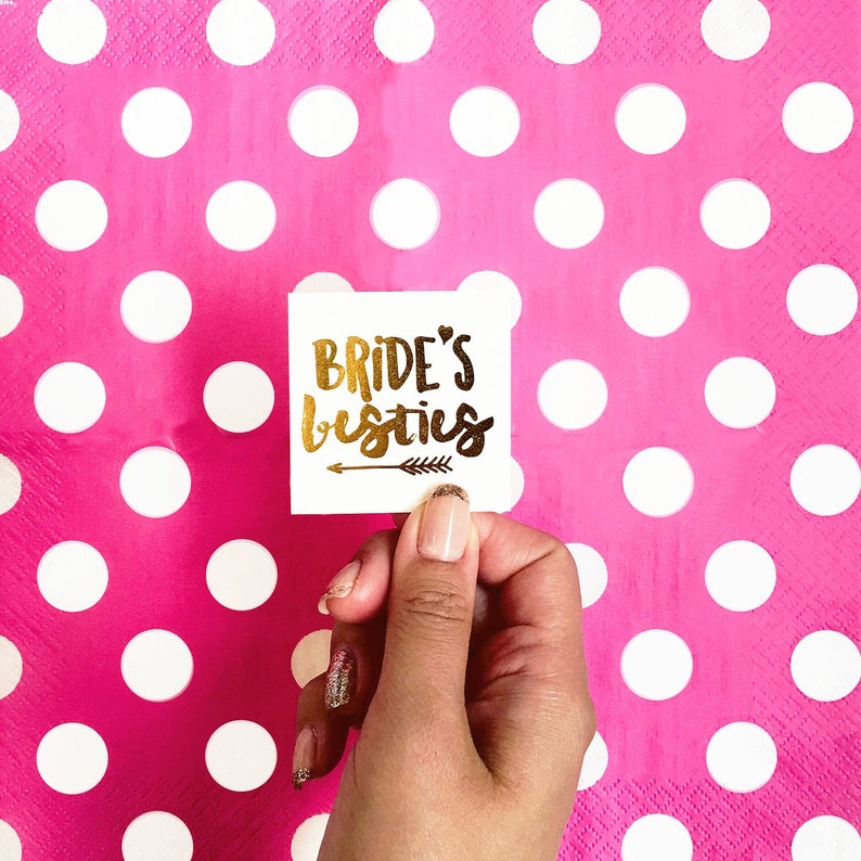 May include: White square temporary tattoo with gold lettering that reads "Bride's besties" with a small gold arrow underneath.