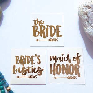 May include: Three gold foil temporary tattoos with arrow designs. The tattoos read "the bride", "bride's besties", and "maid of honor".