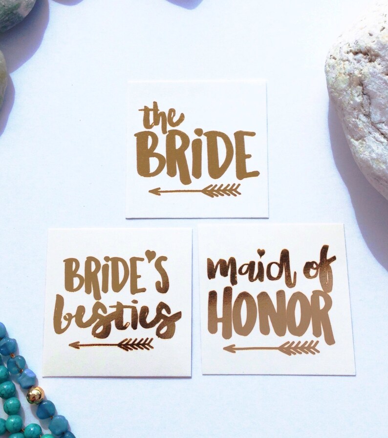 May include: Three gold foil temporary tattoos with arrow designs. The tattoos read "the bride", "bride's besties", and "maid of honor".