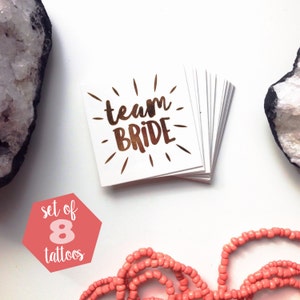 May include: A set of eight gold foil temporary tattoos with the text "team bride" in a script font. The tattoos are on a white background and are arranged in a stack. The text "set of 8 tattoos" is written in white on a pink hexagonal background.