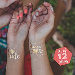 Op de afbeelding: Twee gouden tijdelijke tatoeages op polsen. Eén zegt "bride" met een diamant erboven. De andere zegt "team bride". Een rode cirkel met witte tekst zegt "set of 12 tattoos".