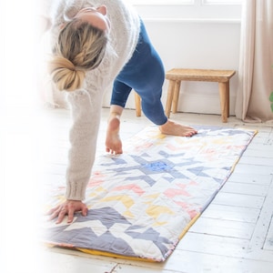 Op de afbeelding: Een vrouw beoefent yoga op een kleurrijke patchwork yoga mat. De mat heeft een geometrisch patroon in tinten blauw, roze, geel en wit. De mat ligt op een witte houten vloer.