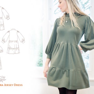 May include: A green jersey dress with a high neck and long sleeves. The dress has a tiered skirt and a relaxed fit. The dress is shown on a model standing in front of a window.