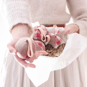 Może przedstawiać: Trzy ręcznie robione świąteczne ozdoby z tkaniny w czerwoną i białą kratę na beżowym tle. Ozdoby mają kształt gwiazdy i kuli. Ozdoby są trzymane przez dłonie osoby.