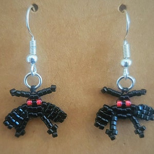 May include: A pair of black beaded earrings with red accents shaped like a spider. The earrings are hanging from silver hooks.