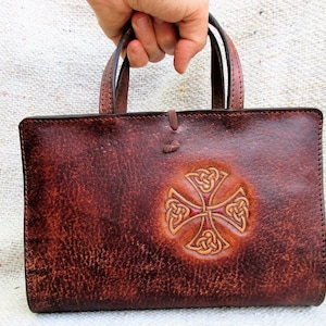 Può includere: Una borsa in pelle marrone con un design a croce di nodo celtico. La borsa ha un'unica maniglia e una chiusura a lembo.