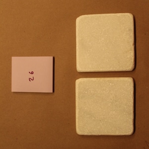 May include: Two white square ceramic coasters with a rough texture. A pink sticky note with the number "26" written in purple is on a brown surface.