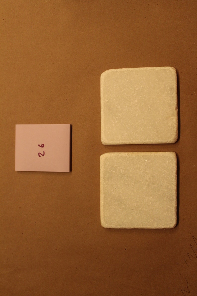 May include: Two white square ceramic coasters with a rough texture. A pink sticky note with the number "26" written in purple is on a brown surface.