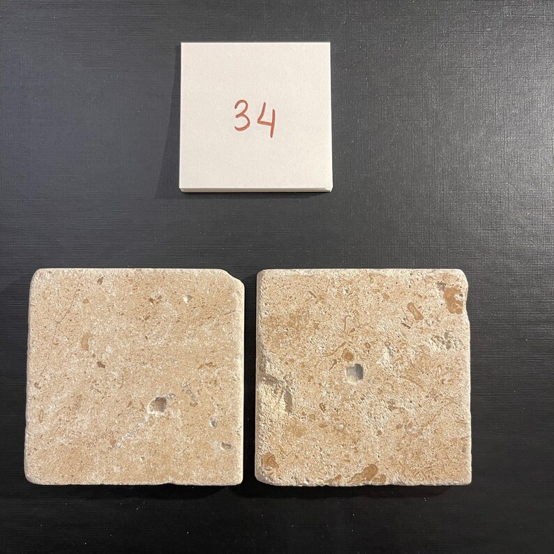 May include: Two beige square tiles with a rough, textured surface. The tiles have a light brown color with small, darker brown flecks throughout. The tiles are arranged side-by-side.