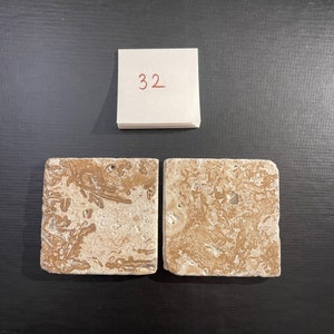 May include: Two square tiles with a light brown and white marbled pattern. The tiles are made of natural stone and have a rough texture. The number 32 is written on a white piece of paper above the tiles.