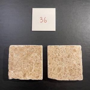 May include: Two beige travertine tile samples with a light brown veining pattern. The tiles are square and have a rough, textured surface. The number 36 is written on a white note card above the tiles.