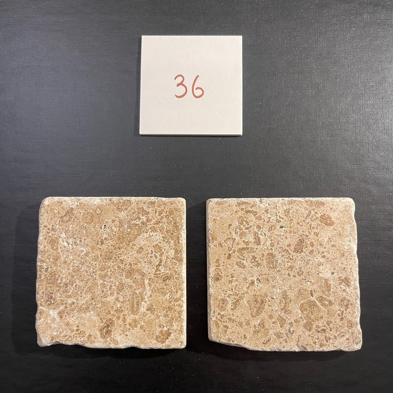 May include: Two beige travertine tile samples with a light brown veining pattern. The tiles are square and have a rough, textured surface. The number 36 is written on a white note card above the tiles.