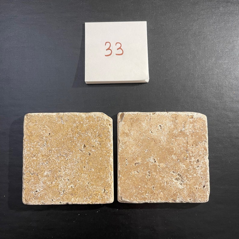 May include: Two square beige travertine tiles with a rough, textured surface. The tiles are arranged side by side.