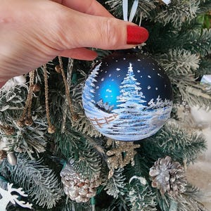 May include: A blue glass Christmas ornament with a winter scene painted on it. The scene includes a snowy forest with a fence and a house in the distance. The ornament is hanging on a Christmas tree.