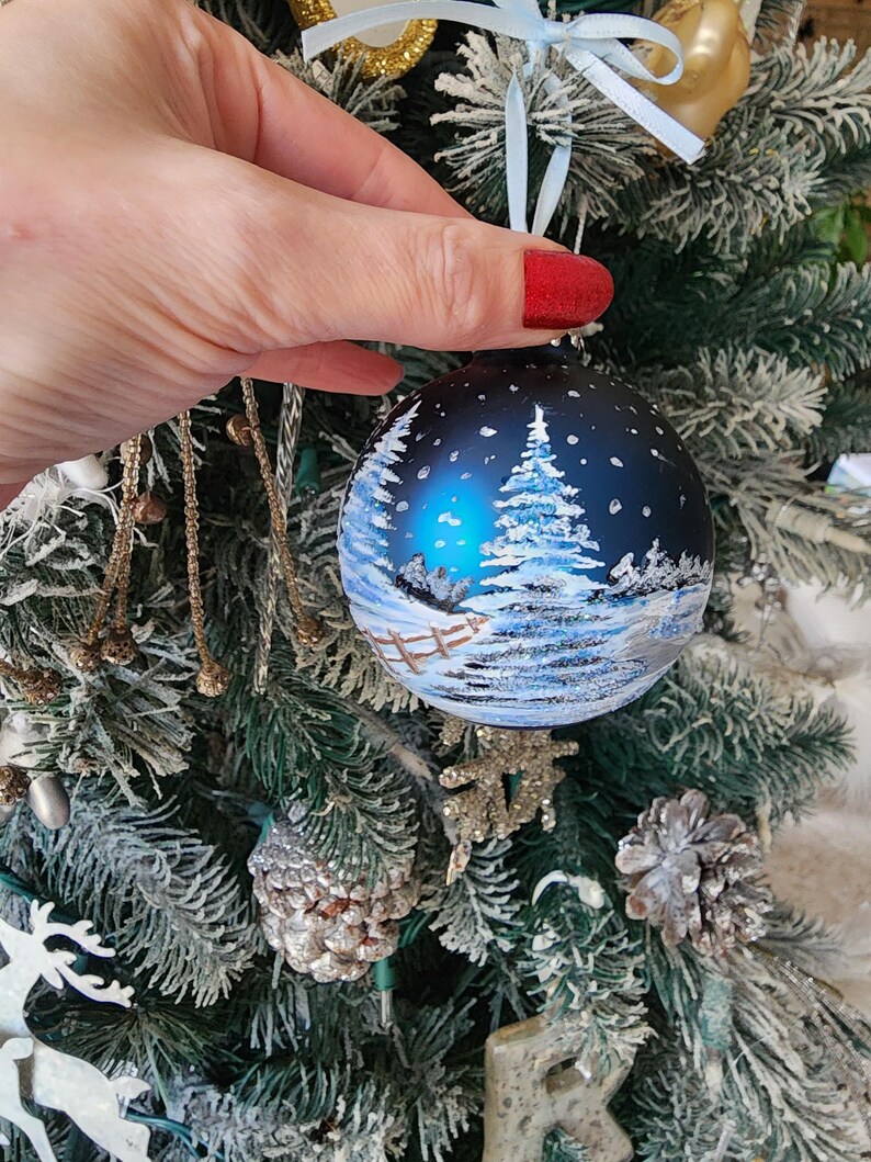 May include: A blue glass Christmas ornament with a winter scene painted on it. The scene includes a snowy forest with a fence and a house in the distance. The ornament is hanging on a Christmas tree.