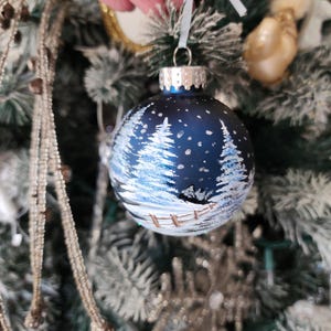 May include: A blue glass Christmas ornament with a winter scene painted on it. The scene includes white trees, a fence, and a snowy background.