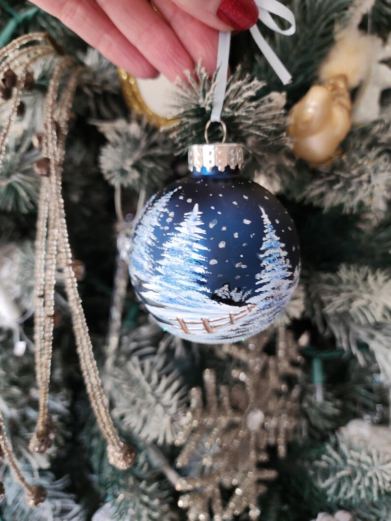 May include: A blue glass Christmas ornament with a winter scene painted on it. The scene includes white trees, a fence, and a snowy background.