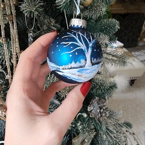 May include: A blue glass Christmas ornament with a white painted winter scene. The ornament features a bare tree with snow on the ground and a snowy sky.