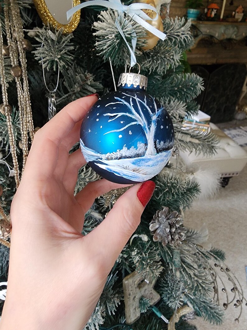 May include: A blue glass Christmas ornament with a white painted winter scene. The ornament features a bare tree with snow on the ground and a snowy sky.