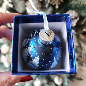 May include: A blue glass Christmas ornament with a white winter scene design. The ornament is in a blue box with a white ribbon.