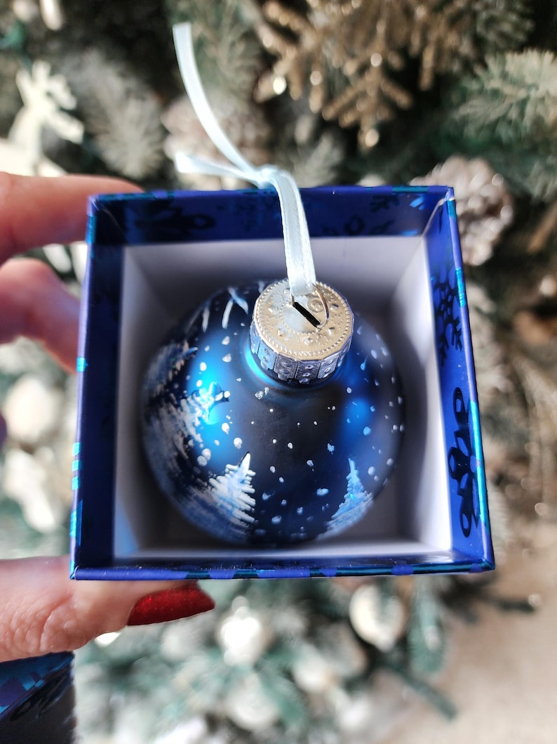 May include: A blue glass Christmas ornament with a white winter scene design. The ornament is in a blue box with a white ribbon.