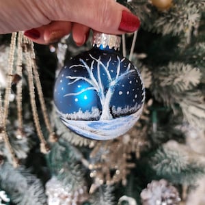 May include: A blue glass Christmas ornament with a white painted winter scene of a tree and snow. The ornament has a silver top and is hanging on a Christmas tree.