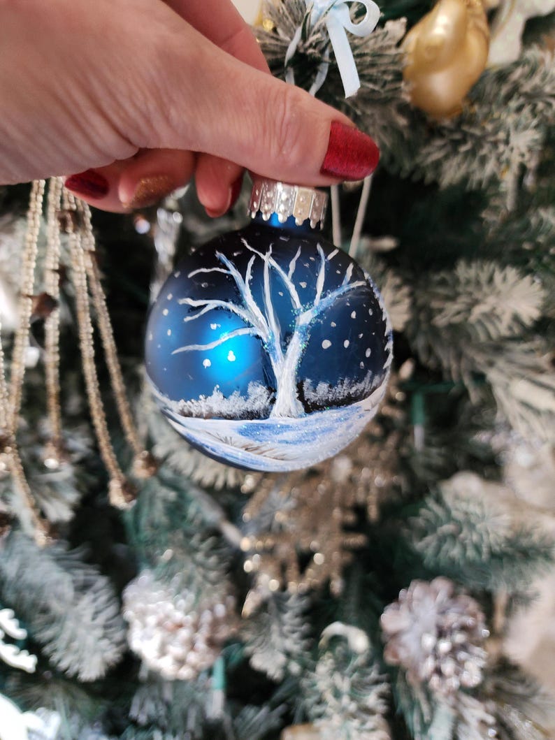 May include: A blue glass Christmas ornament with a white painted winter scene of a tree and snow. The ornament has a silver top and is hanging on a Christmas tree.