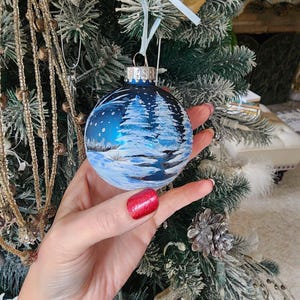 May include: A blue glass Christmas ornament with a winter scene painted on it. The scene includes snow-covered trees, a snowy landscape, and a blue sky with snowflakes.