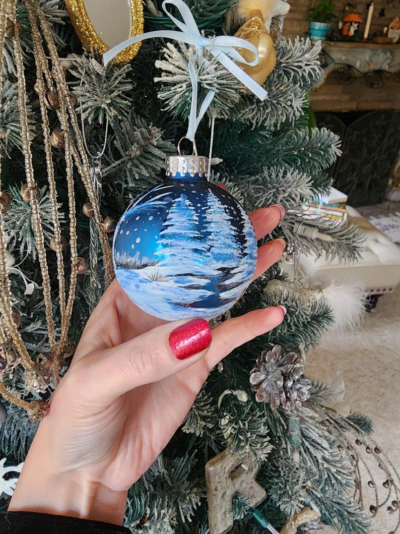 May include: A blue glass Christmas ornament with a winter scene painted on it. The scene includes snow-covered trees, a snowy landscape, and a blue sky with snowflakes.