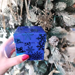 May include: A small blue gift box with a snowflake pattern. The box is being held by a hand with red nail polish.