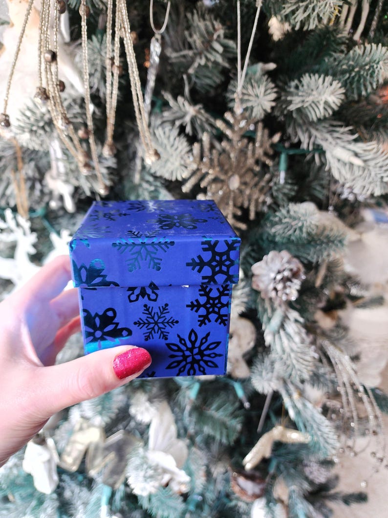 May include: A small blue gift box with a snowflake pattern. The box is being held by a hand with red nail polish.