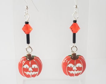 May include: A pair of Halloween-themed earrings. Each earring features a red pumpkin charm with a silver face, suspended from a silver hook. Black and orange beads add detail to the design. The earrings are displayed on a clear stand.