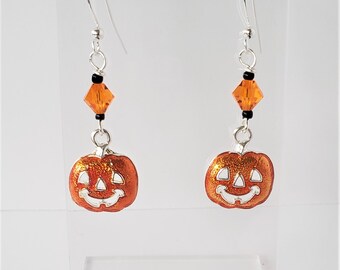 May include: A pair of Halloween-themed earrings. Each earring features a smiling orange pumpkin charm with a white face, suspended from a silver hook. The design includes an orange crystal and black beads.