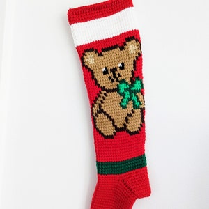 May include: A red Christmas stocking with a brown teddy bear design. The stocking has a white band at the top, a green stripe near the bottom, and a loop for hanging. The teddy bear has a green bow.