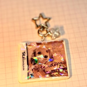 May include: A square keychain featuring a miniature replica of the Nintendogs Dachshund & Friends Nintendo DS game case. The surface has a holographic finish with scattered light-reflecting shards. It includes a silver-toned star-shaped clasp.