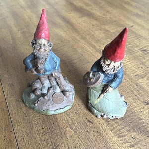 May include: Two garden gnome figurines. One gnome is seated, wearing a red hat and blue jacket, holding a small object. The other gnome is painting, wearing a red hat and blue jacket, with a green leaf base.