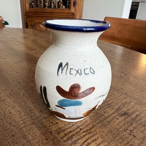 May include: A ceramic vase with a cream-colored body and a dark blue rim. The word "Mexico" is written in black script. The vase features a painted design of a sombrero and other decorative elements in brown, blue, and green.