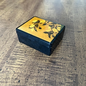 May include: A small black wooden box with a floral design on the lid. The lid is decorated with yellow flowers and black leaves on a gold background.