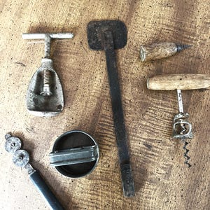 May include: An assortment of vintage bar tools, including a metal corkscrew, a bottle opener, and a can opener. The tools are made of metal and wood, with signs of wear and age. The items are arranged on a wooden surface.