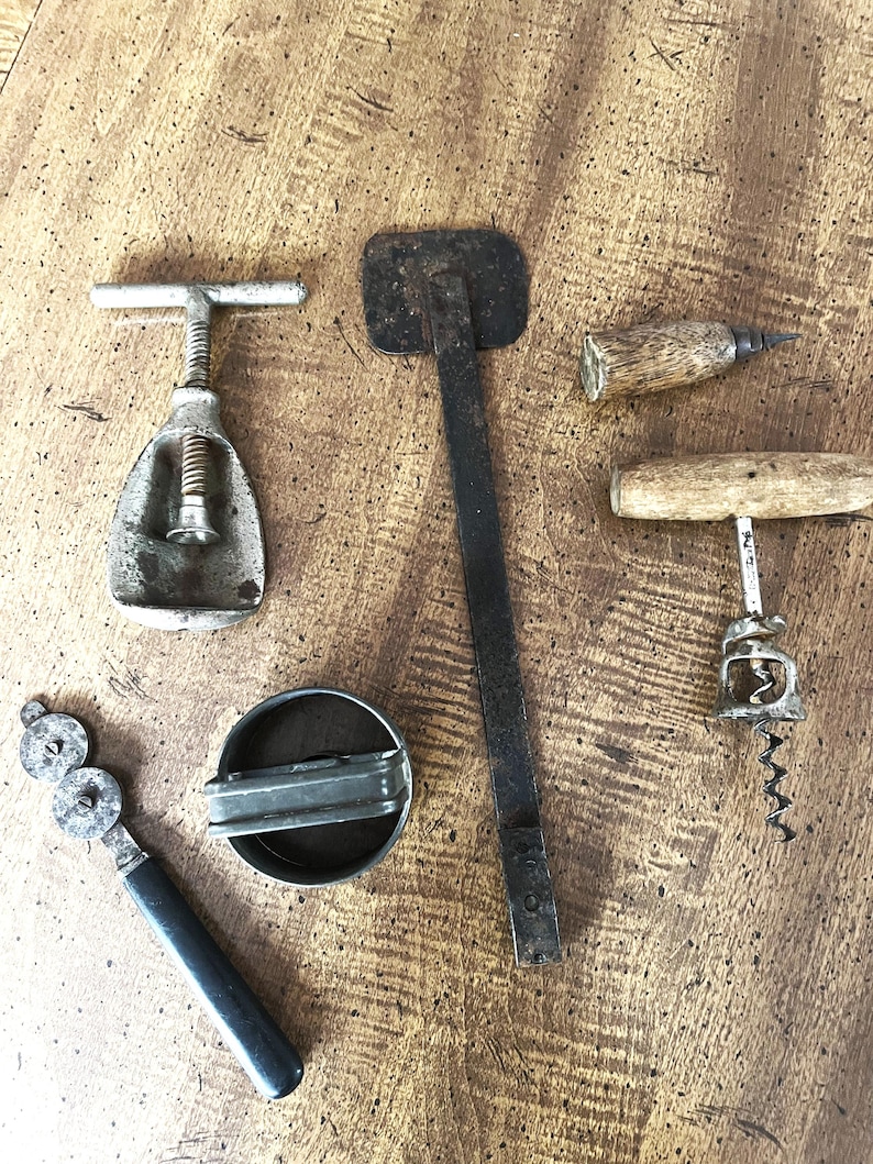 May include: An assortment of vintage bar tools, including a metal corkscrew, a bottle opener, and a can opener. The tools are made of metal and wood, with signs of wear and age. The items are arranged on a wooden surface.