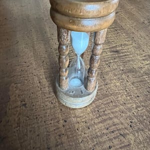 K&ouml;nnte beinhalten: Eine h&ouml;lzerne Sanduhr mit hellem Sand. Die Sanduhr wird von einem Holzrahmen mit dekorativen gedrehten S&auml;ulen getragen. Das Holz hat eine warme, nat&uuml;rliche Oberfl&auml;che. Ein klassisches Zeitmessger&auml;t.