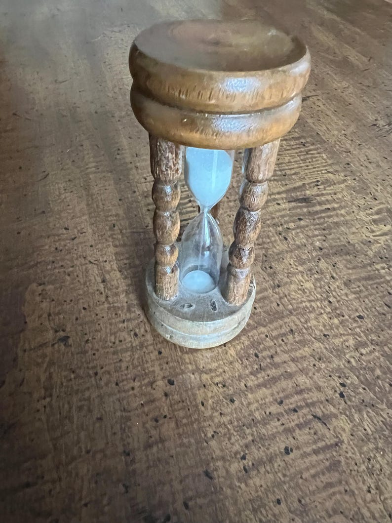 K&ouml;nnte beinhalten: Eine h&ouml;lzerne Sanduhr mit hellem Sand. Die Sanduhr wird von einem Holzrahmen mit dekorativen gedrehten S&auml;ulen getragen. Das Holz hat eine warme, nat&uuml;rliche Oberfl&auml;che. Ein klassisches Zeitmessger&auml;t.