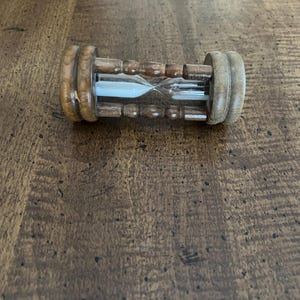 K&ouml;nnte beinhalten: Eine h&ouml;lzerne Sanduhr mit hellbraunen Holzendkappen und einer dunkelbraunen Holzmitte. Die Glaskammer enth&auml;lt wei&szlig;en Sand. Die Sanduhr liegt auf einer dunkelbraunen Holzoberfl&auml;che.