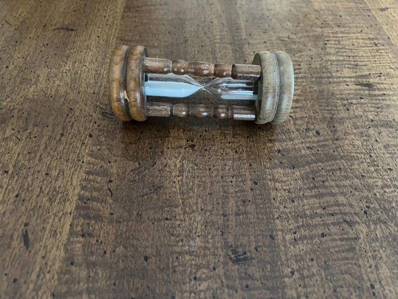 K&ouml;nnte beinhalten: Eine h&ouml;lzerne Sanduhr mit hellbraunen Holzendkappen und einer dunkelbraunen Holzmitte. Die Glaskammer enth&auml;lt wei&szlig;en Sand. Die Sanduhr liegt auf einer dunkelbraunen Holzoberfl&auml;che.