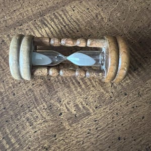 K&ouml;nnte beinhalten: Eine h&ouml;lzerne Sanduhr mit hellem Sand in einer durchsichtigen Glaskugel. Die Sanduhr hat einen hellbraunen Holzrahmen mit abgerundeten Enden und einer schmalen Mitte. Ein klassisches Zeitmessger&auml;t.