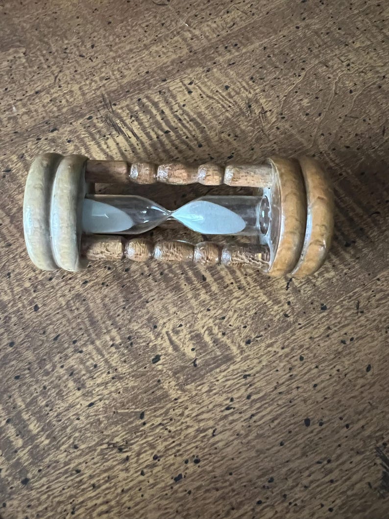 K&ouml;nnte beinhalten: Eine h&ouml;lzerne Sanduhr mit hellem Sand in einer durchsichtigen Glaskugel. Die Sanduhr hat einen hellbraunen Holzrahmen mit abgerundeten Enden und einer schmalen Mitte. Ein klassisches Zeitmessger&auml;t.