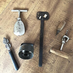 May include: An assortment of vintage kitchen tools, including a can opener, corkscrews, and a metal pastry cutter, arranged on a wooden surface. The tools have a rustic, well-used appearance, with metal and wooden handles.