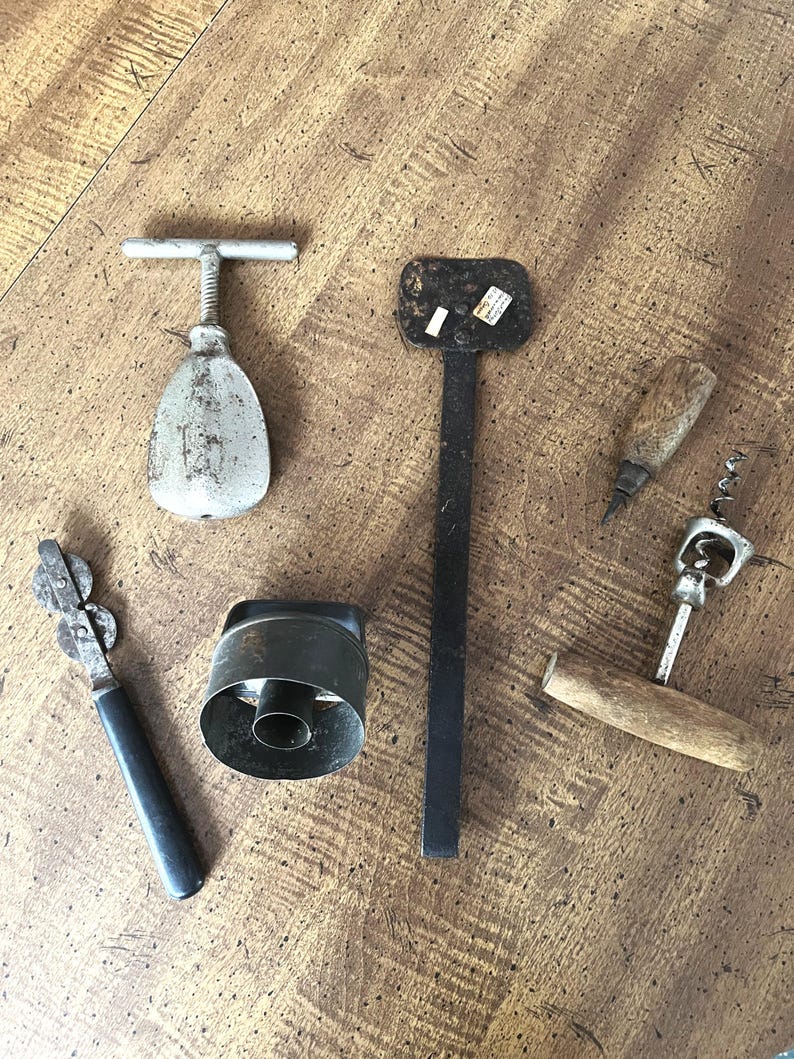 May include: An assortment of vintage kitchen tools, including a can opener, corkscrews, and a metal pastry cutter, arranged on a wooden surface. The tools have a rustic, well-used appearance, with metal and wooden handles.